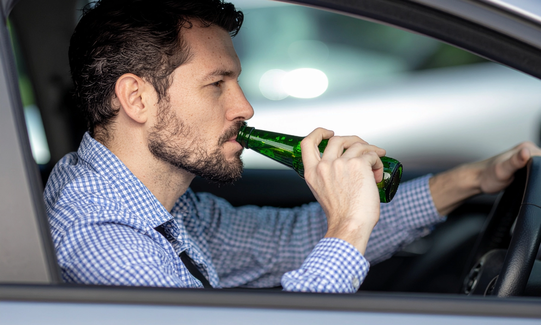 A man with a beard sits in the driver’s seat of a car, wearing a blue checkered shirt, drinking from a green bottle while holding the steering wheel—an image that might concern any Las Vegas personal injury lawyer.